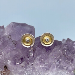 Zwei Ohrstecker in Silber und Gelbgold mit Brillant liegen auf einem Amethyst vor blauem Hintergrund.