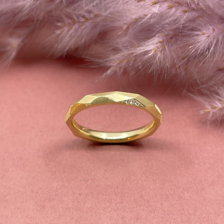 Ring "Frances" - 585 Gelbgold mit großen Facetten Trauring Frances steht schräg auf einem pinken Untergrund, die Diamanten und gefeilten Facetten sind schön sichtbar.